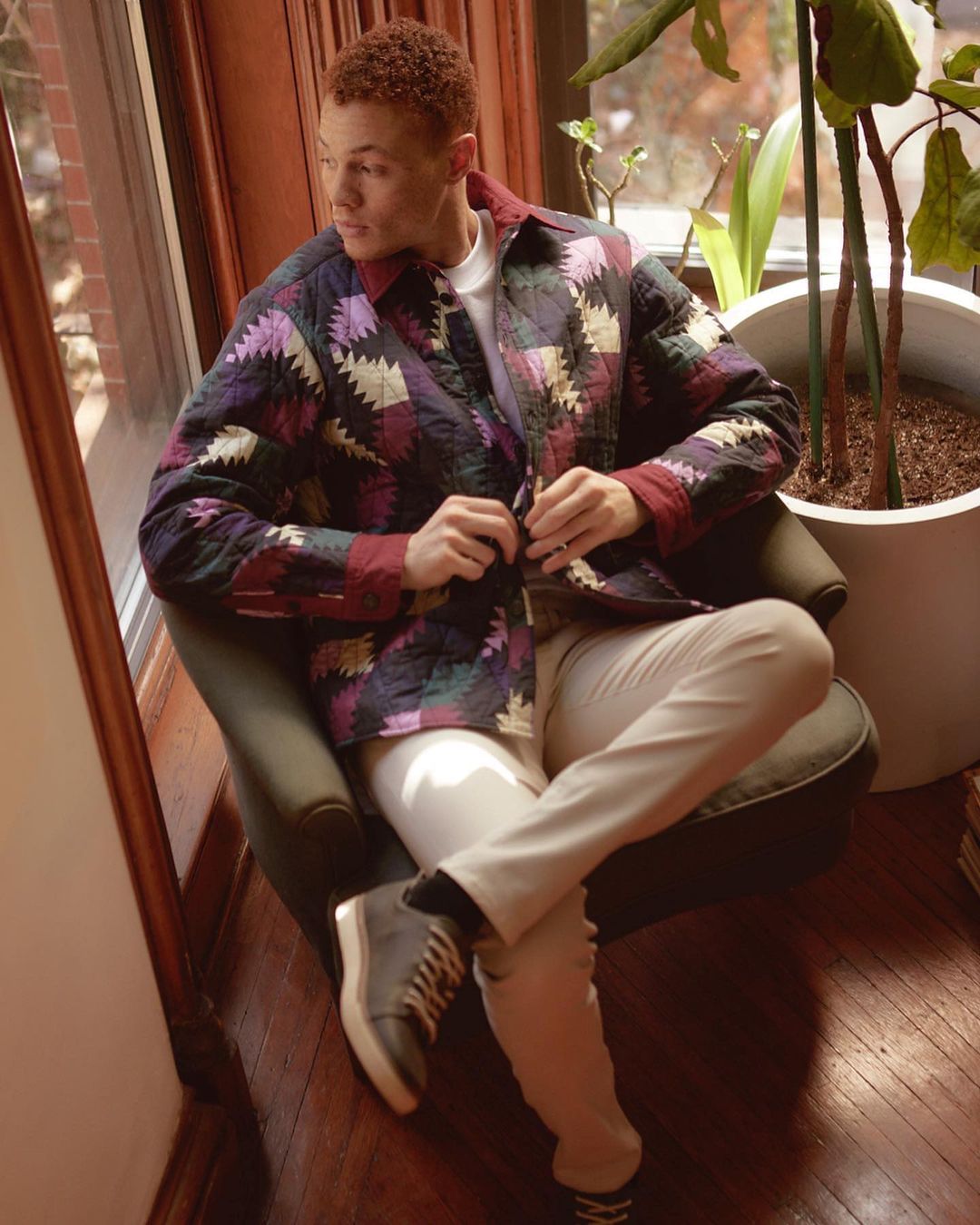 A red-haired man in a patchwork jacket sits near a window. His legs are crossed, and he is wearing a pair of grey shoes and light pants.