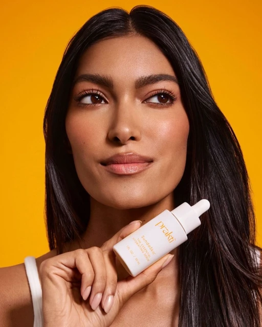 A tan-skinned woman with dark hair smiles softly, holding a bottle of skincare serum against a bright yellow background in a beauty product shot.