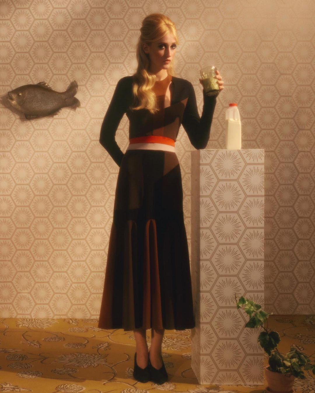 Elegant woman in brown dress holds green smoothie. Milk carton on geometric stand. Fish art and patterned wallpaper backdrop.