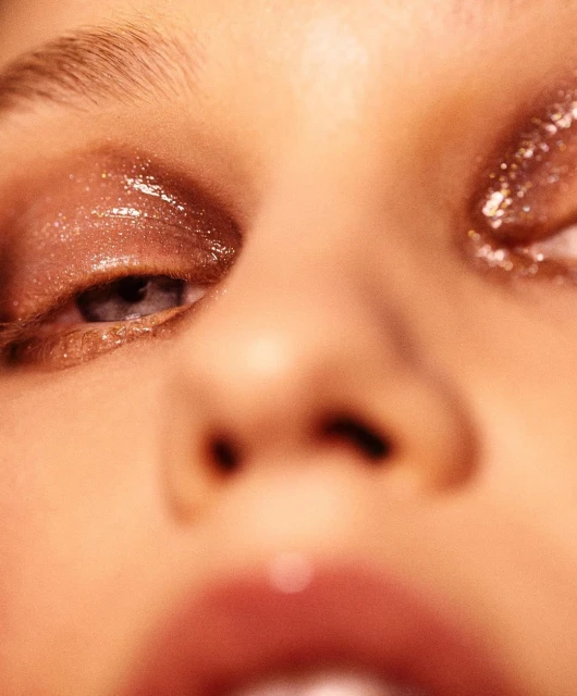 Close-up of a woman's face features glossy eye makeup. The focus is on her eyes and the shimmering effect of the makeup.