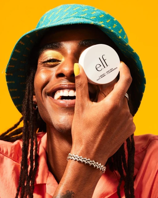 Person holding e.l.f. putty primer in front of face, wearing blue bucket hat, peach shirt, and silver bracelet against a yellow backdrop.
