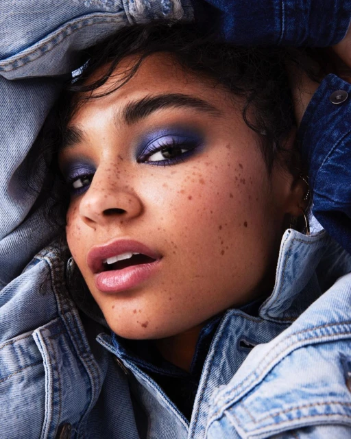 Close-up portrait of a young adult female with blue eyeshadow and freckles wearing a light blue denim jacket.