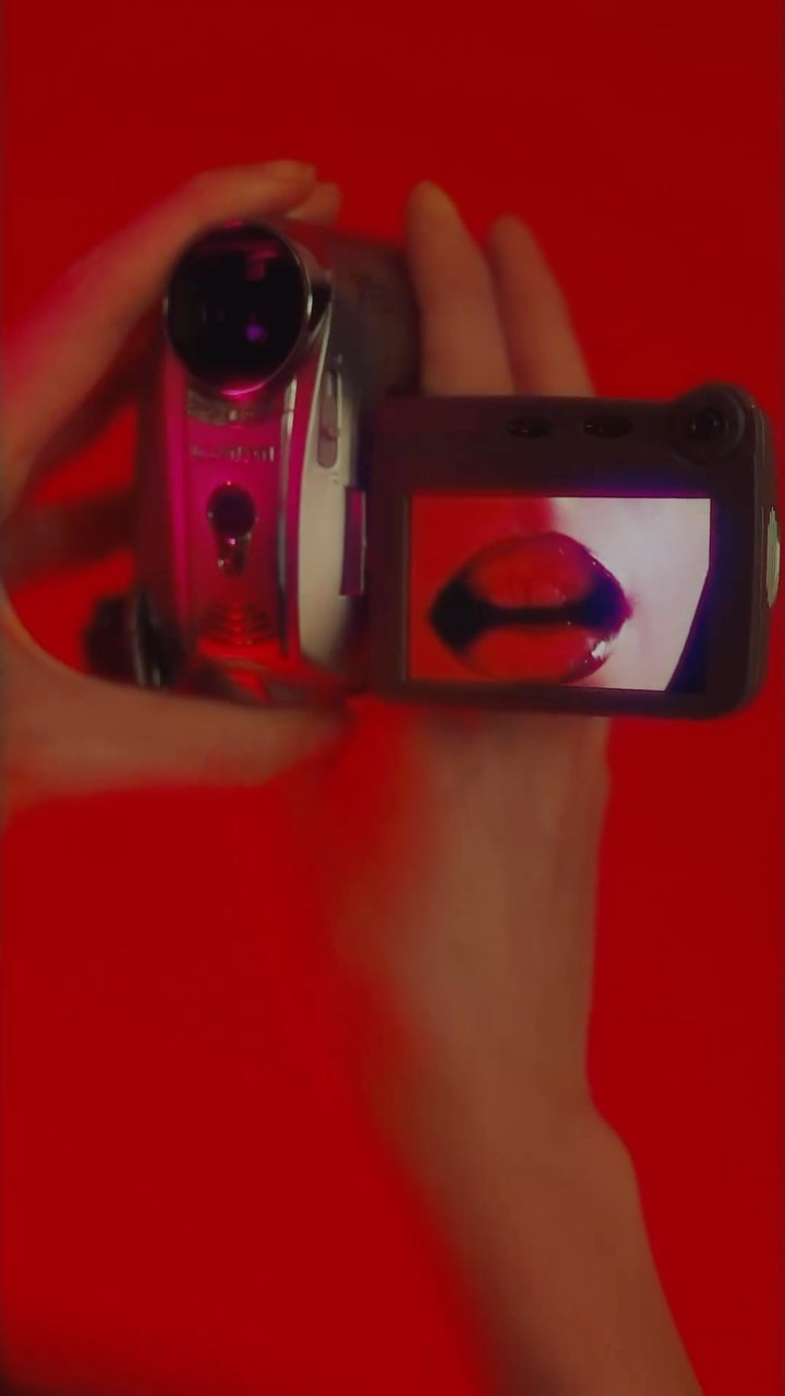 Hands hold a camcorder, the screen displaying a red-lipped face. The scene is set against a solid red background.