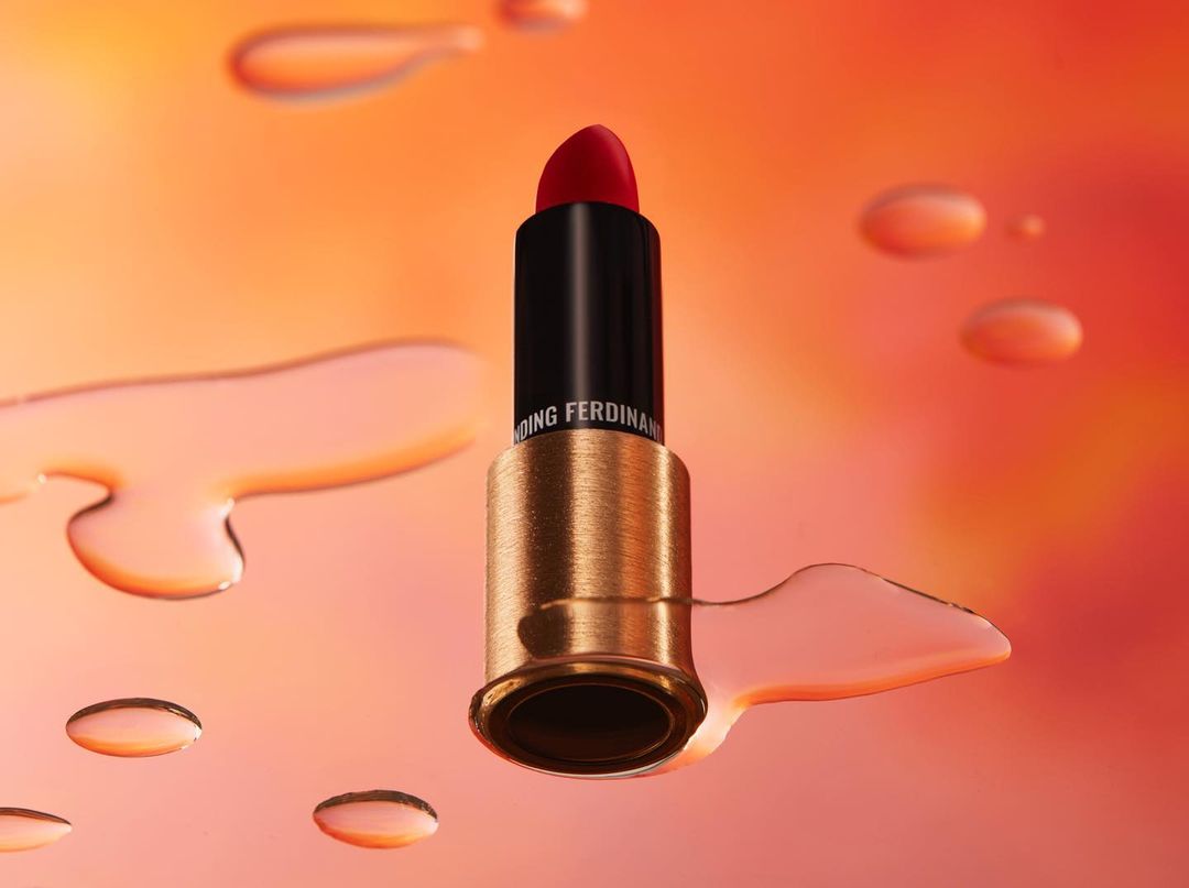 Elevated studio shot of red lipstick in black and gold tube surrounded by translucent water droplets on an orange gradient background.