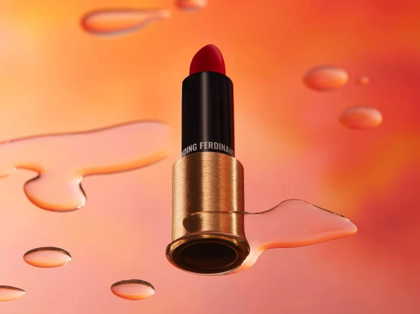 Elevated studio shot of red lipstick in black and gold tube surrounded by translucent water droplets on an orange gradient background.
