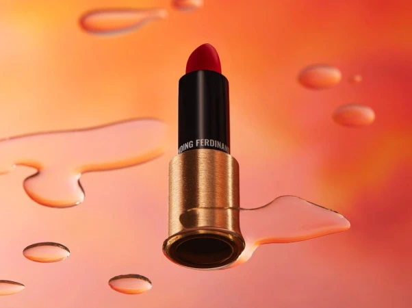Elevated studio shot of red lipstick in black and gold tube surrounded by translucent water droplets on an orange gradient background.