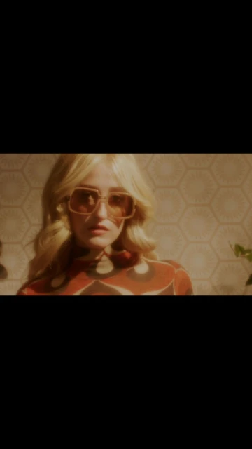 Adult female with blonde hair wearing large, tinted glasses and a patterned orange blouse poses in front of a patterned wall.