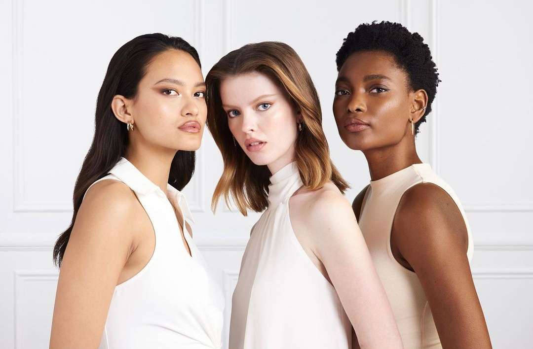 Close-up studio shot of three diverse women, showcasing their natural makeup and hairstyles. The women are wearing elegant, sleeveless tops.