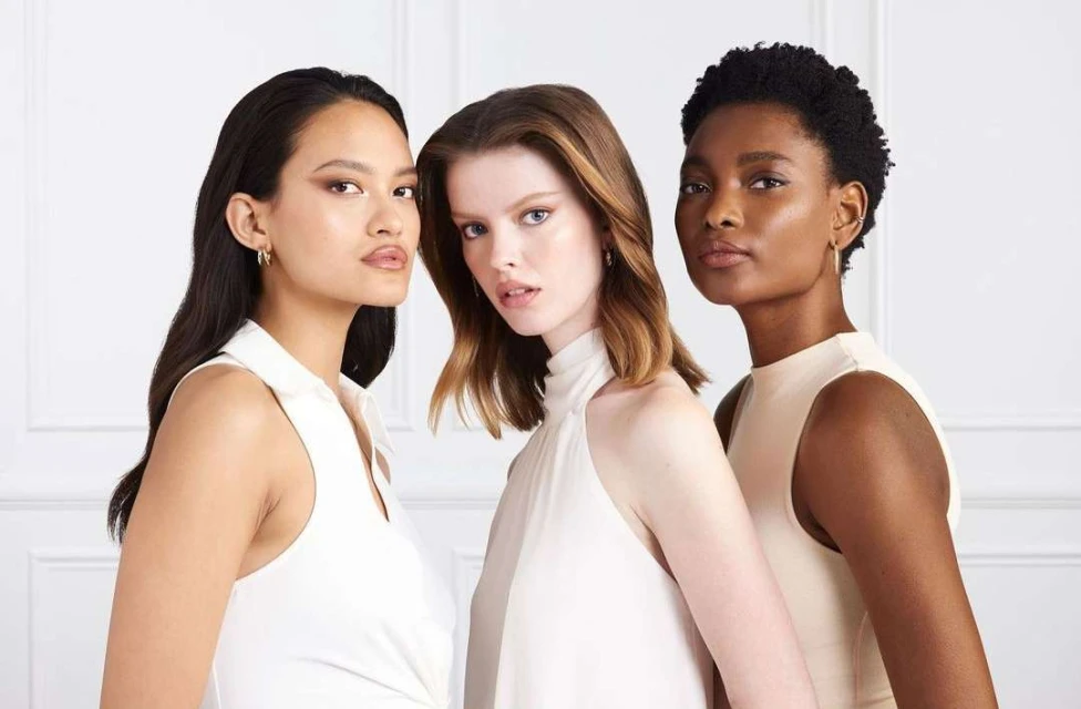 Close-up studio shot of three diverse women, showcasing their natural makeup and hairstyles. The women are wearing elegant, sleeveless tops.