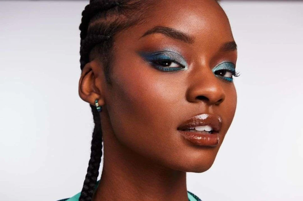 Close-up portrait of a dark-skinned woman with creative teal and blue eye makeup, glossy lips, and braided hair against a white backdrop.