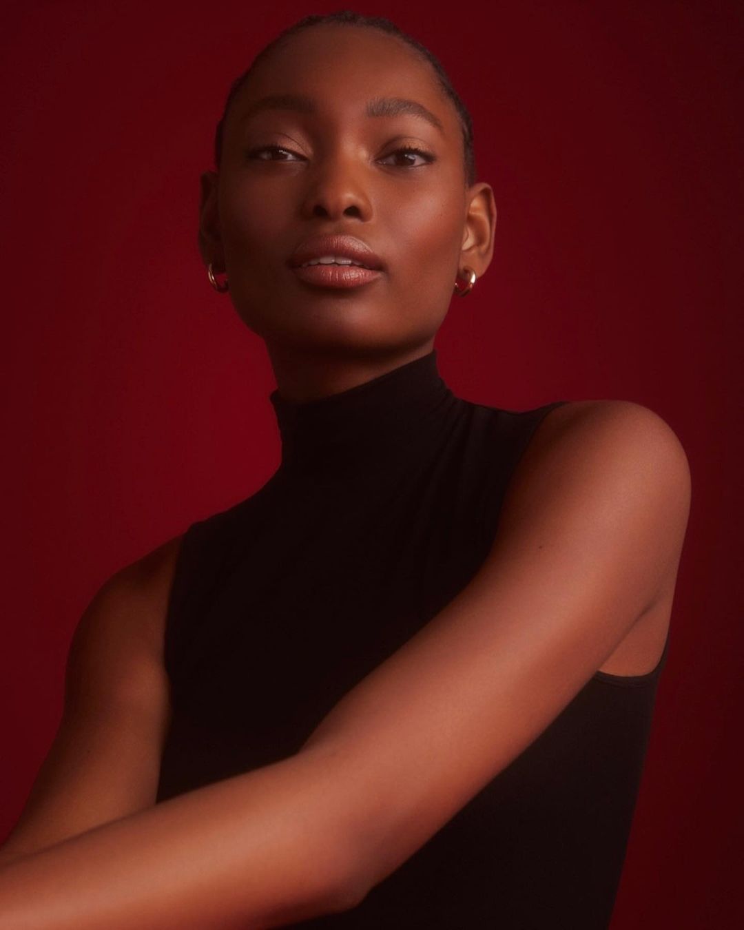 Close-up studio portrait of a Black woman in a sleeveless turtleneck against a red background, lit with soft, diffused light.