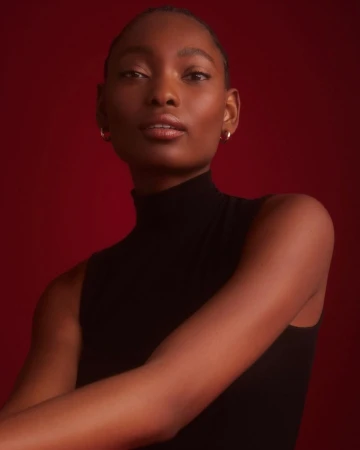 Close-up studio portrait of a Black woman in a sleeveless turtleneck against a red background, lit with soft, diffused light.
