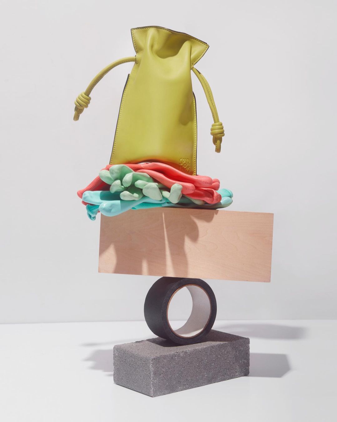 Studio shot of a chartreuse drawstring purse atop a stack of colored objects, including a square, tape roll, and concrete block.