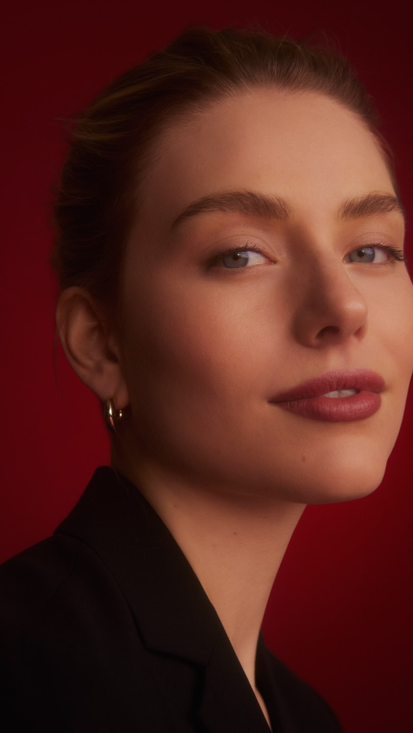 Close-up studio shot of a woman with fair skin, wearing a black blazer and hoop earrings against a solid red backdrop, beauty concept.