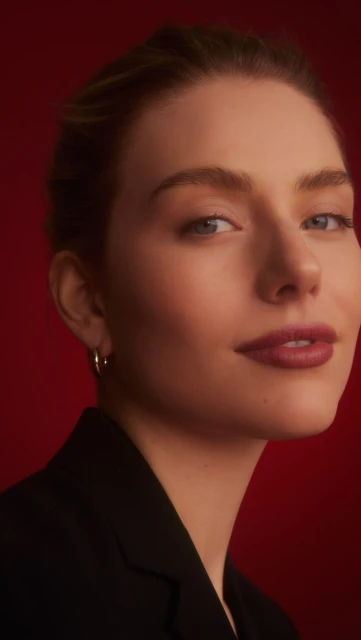 Close-up studio shot of a woman with fair skin, wearing a black blazer and hoop earrings against a solid red backdrop, beauty concept.