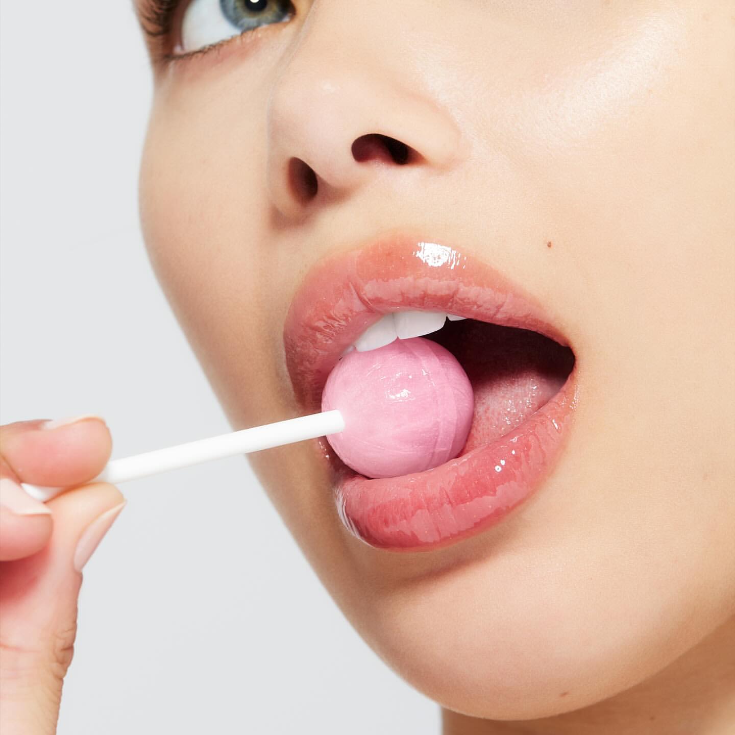 Close-up of a young adult female holding a pink lollipop in her mouth, showcasing glossy lips and bright, clean skin against a plain backdrop.
