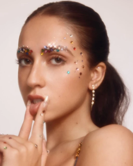 Close-up of a woman with jewel-encrusted makeup around her eyes, lips slightly parted, posing against a plain white background.