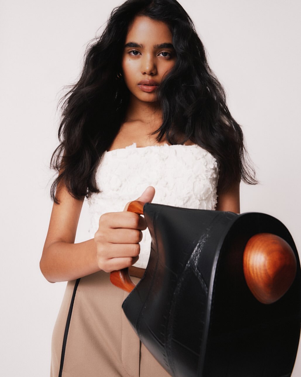 Brunette woman holds a unique black bag with a wooden handle. She poses in a white textured top and tan high-waisted pants in a studio.