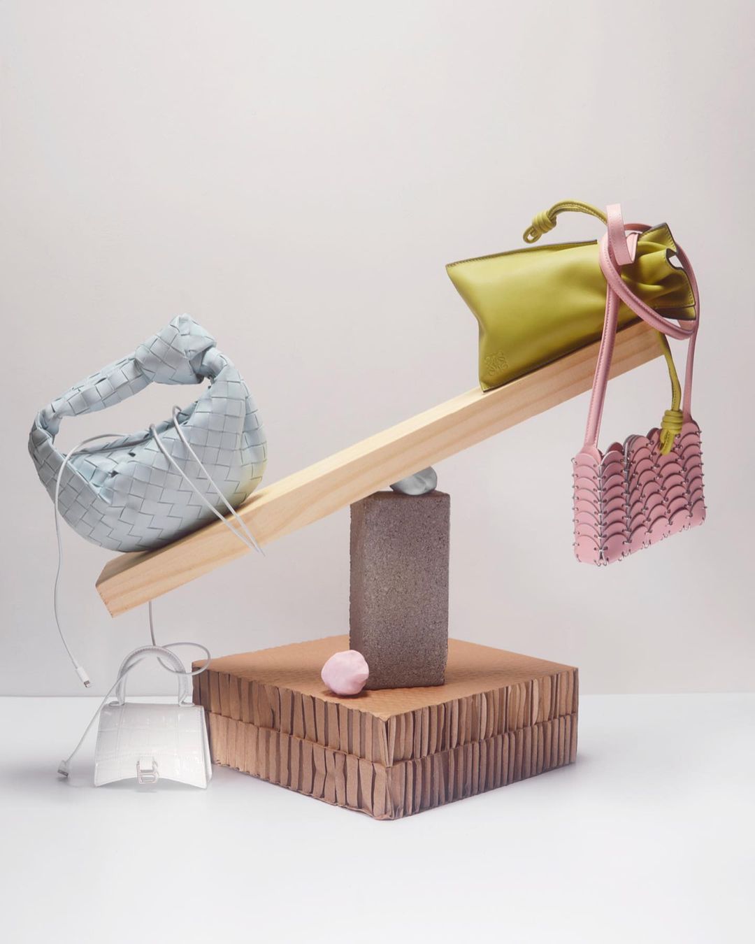 Still life product shot featuring a seesaw balanced with miniature handbags in green, light blue, and pink against a white background.