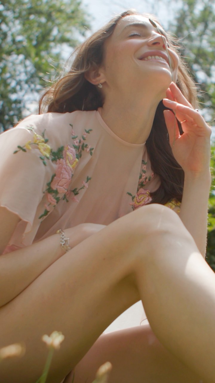 Smiling woman in a light pink embroidered top sits in a grassy outdoor setting with trees and bright sunlight overhead.