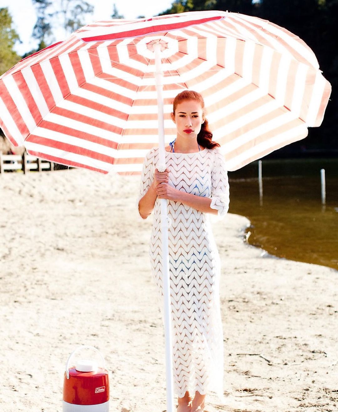 A woman in a white dress stands on a sandy beach holding a red and white striped parasol, with a cooler and lake in the backdrop.