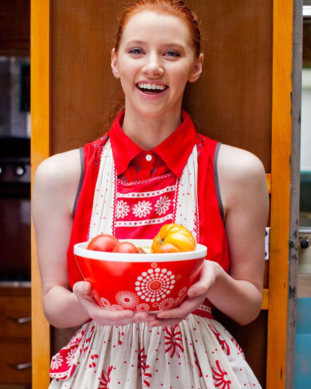 Cheerful redhead woman in a red-and-white dress holds a bowl of tomatoes in a kitchen setting, smiling brightly at the camera.