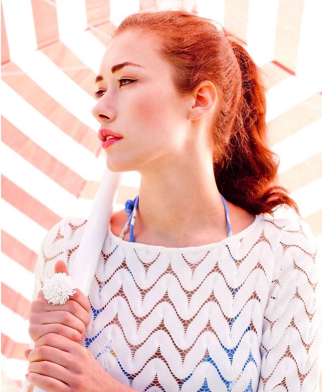 Portrait of a red-haired woman in a white knit top holding a white umbrella, photographed in bright, diffused lighting.