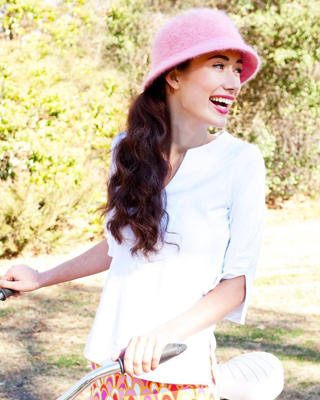 Smiling woman in pink hat stands outdoors holding a bicycle. She wears a white blouse and colourful patterned skirt. Sunny daylight.