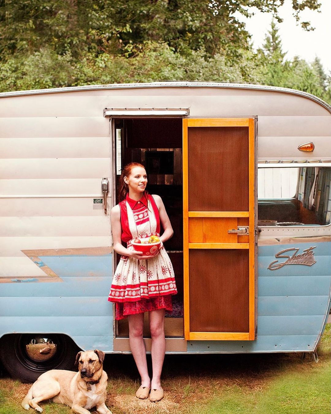 Young woman in red and white dress stands in front of vintage camper with a dog, holding bowl with yellow fruit in a grassy outdoor setting.
