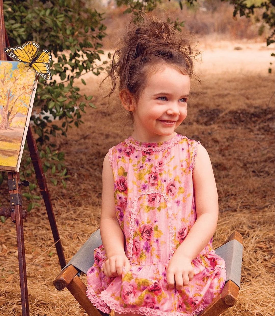 A young girl with curly hair sits on a director's chair next to an easel with a butterfly perched on an art piece in a wooded outdoor setting.