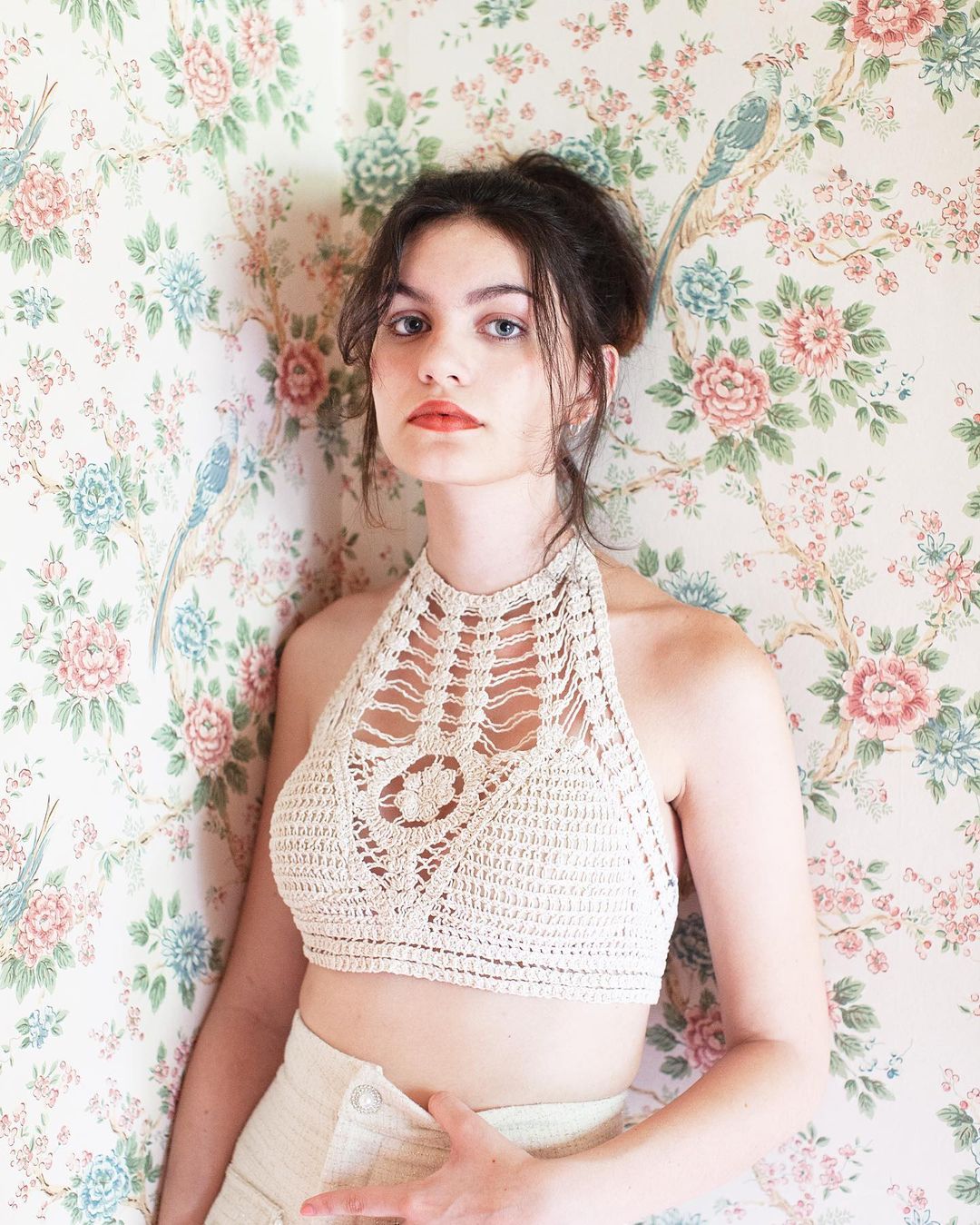 Young woman in a crochet halter top poses against a vintage floral wallpaper, creating a fashion and portrait image with natural light.
