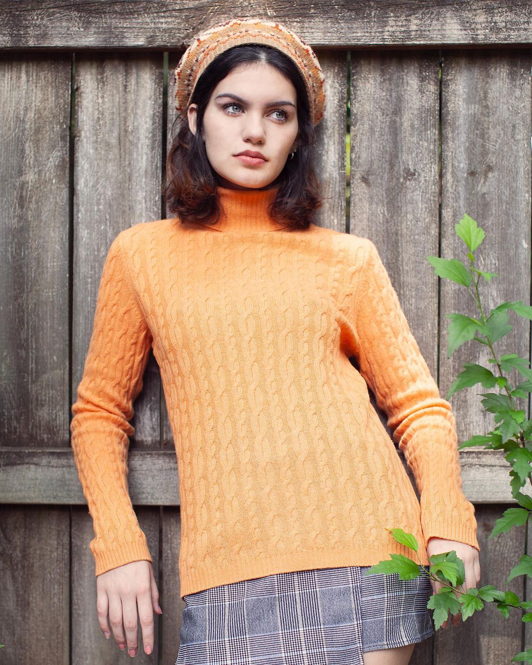 Young adult woman in an orange turtleneck sweater and plaid skirt poses outdoors near a weathered wood fence and green foliage.