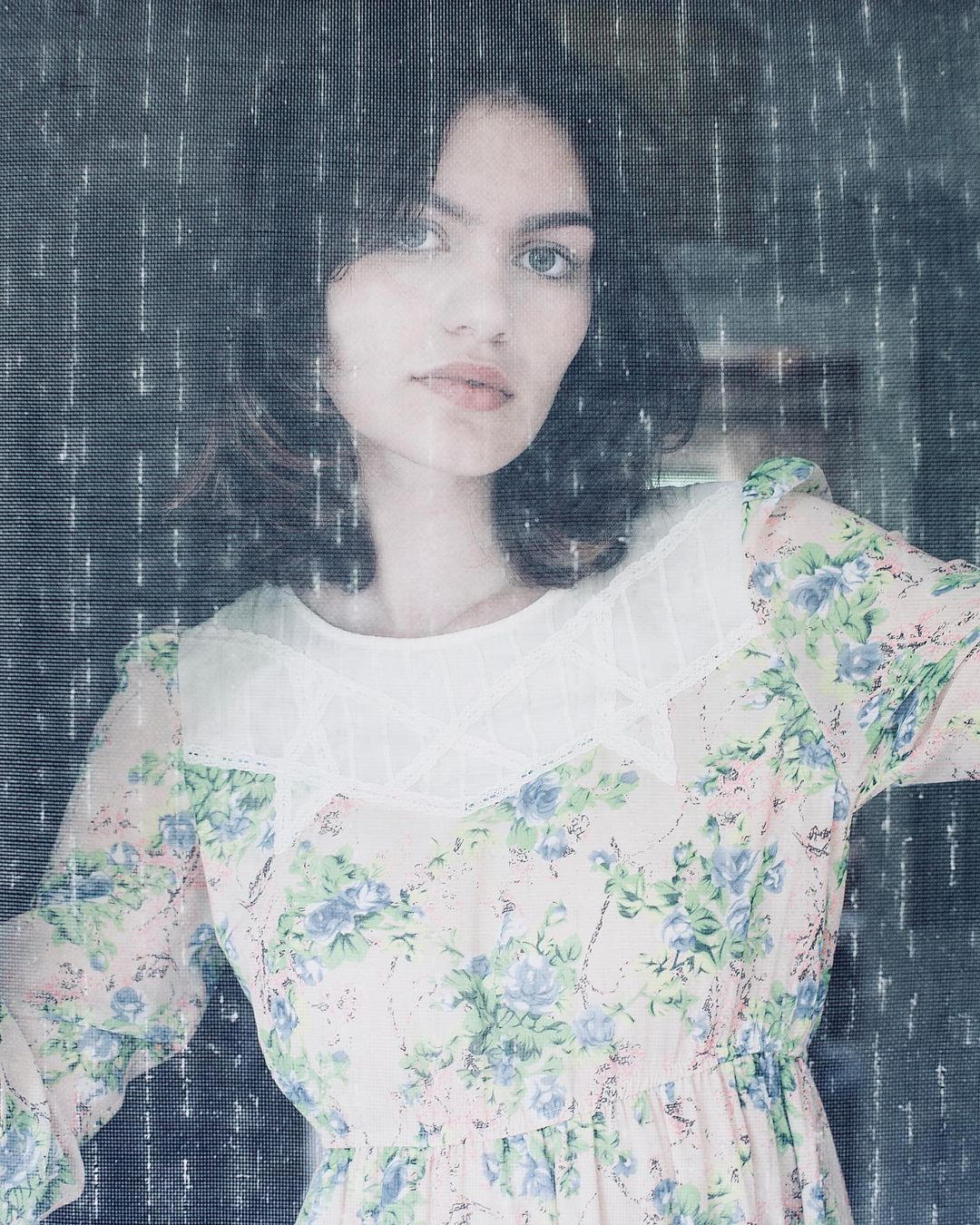 Portrait of a young woman with brown hair and light skin wearing a floral dress with a lace collar against a grey patterned backdrop.