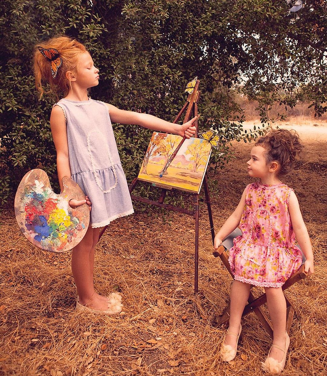 Two young girls are in a grassy outdoor setting, one standing with a paint palette and the other sitting, next to a painting of trees.