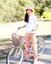 Woman in white top and pink patterned trousers holding a pink bicycle with a flower-filled basket