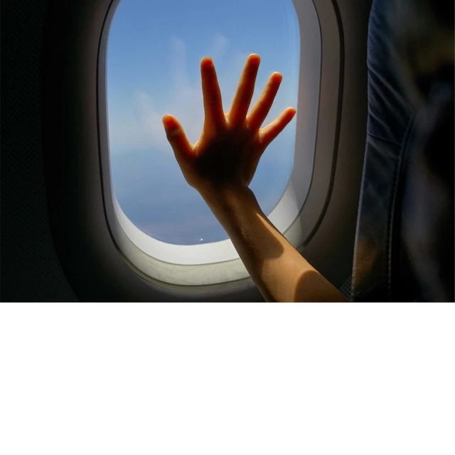 Child's hand pressed against an airplane window during flight, viewing blue sky and clouds through the glass.