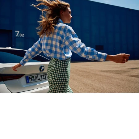 Woman in blue and white checkered shirt and green skirt walks by a white BMW against a blue building backdrop on a sunny day.