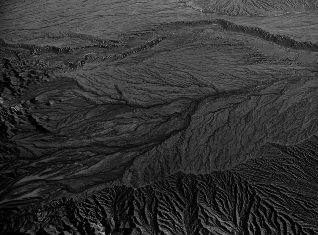 Monochrome aerial view of a textured, mountainous landscape featuring dry terrain with patterns of erosion and deep ravines.