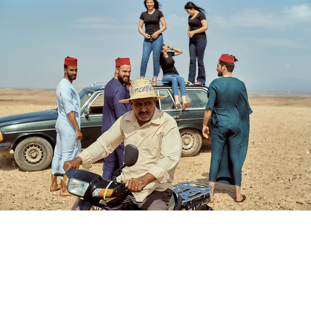 Group of six people in a desert-like setting; three women stand on a vintage Mercedes-Benz, while three men pose near, one on a scooter.
