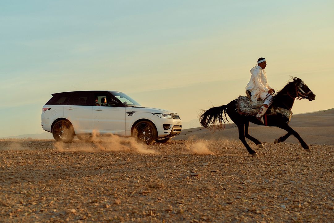 Horseback rider in traditional garb racing a white Range Rover across a desert landscape at sunset with dynamic dust trails.