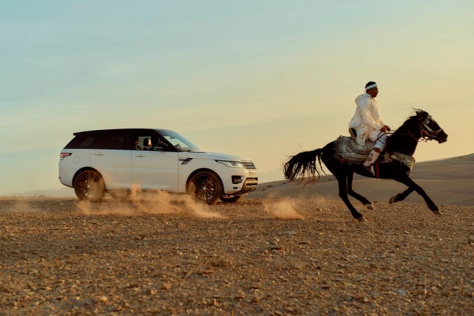 Horseback rider in traditional garb racing a white Range Rover across a desert landscape at sunset with dynamic dust trails.