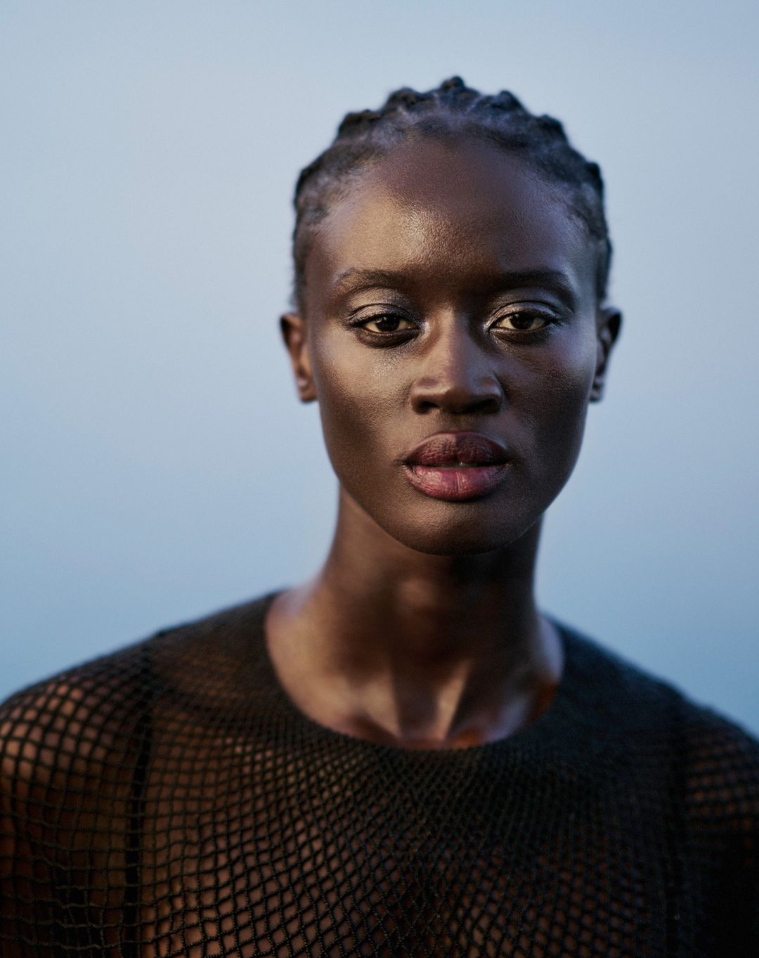 Close-up portrait of a Black woman with braided hair and a dark mesh top, against a light blue backdrop, showcasing beauty and fashion.