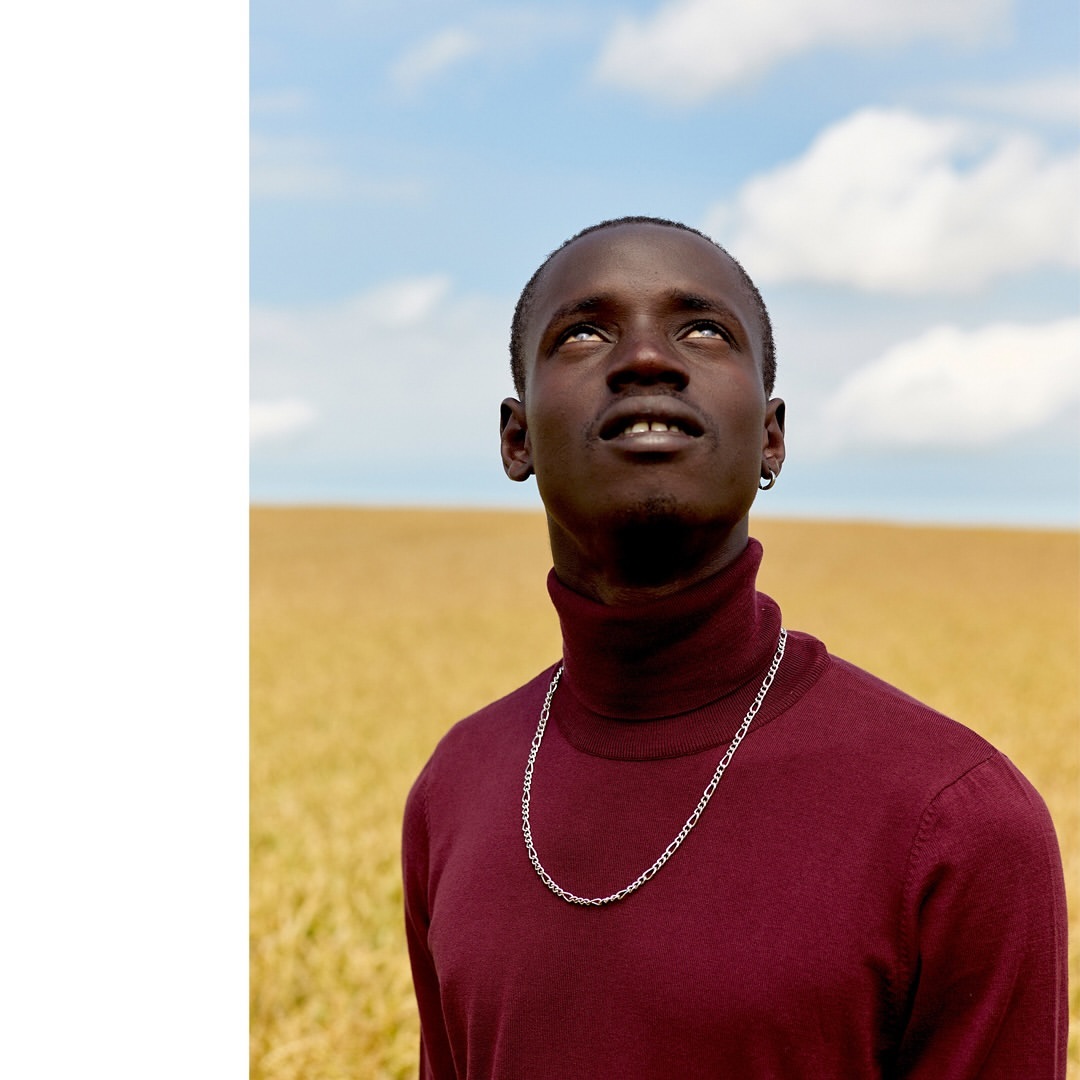 Young adult man with dark skin looks upward in an outdoor field. He wears a burgundy turtleneck and silver chain against a blue sky backdrop.