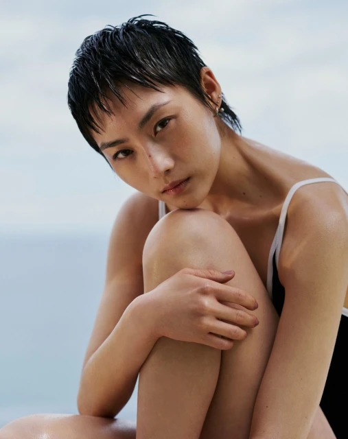 Woman in black one-piece swimsuit poses with hands clasped around knees against blurred water background in beauty-style portrait.