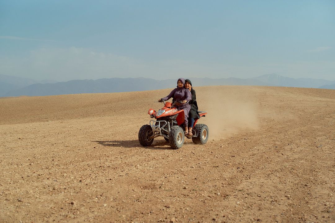 Two adult females ride a red ATV across a tan desert landscape under a clear blue sky, with distant mountains in the background.