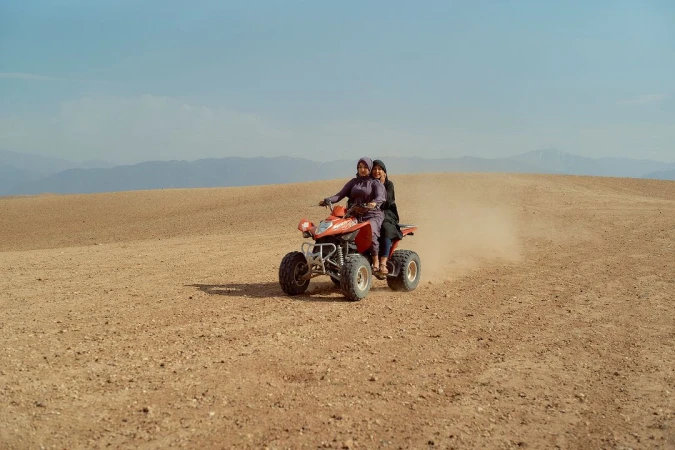 Two adult females ride a red ATV across a tan desert landscape under a clear blue sky, with distant mountains in the background.