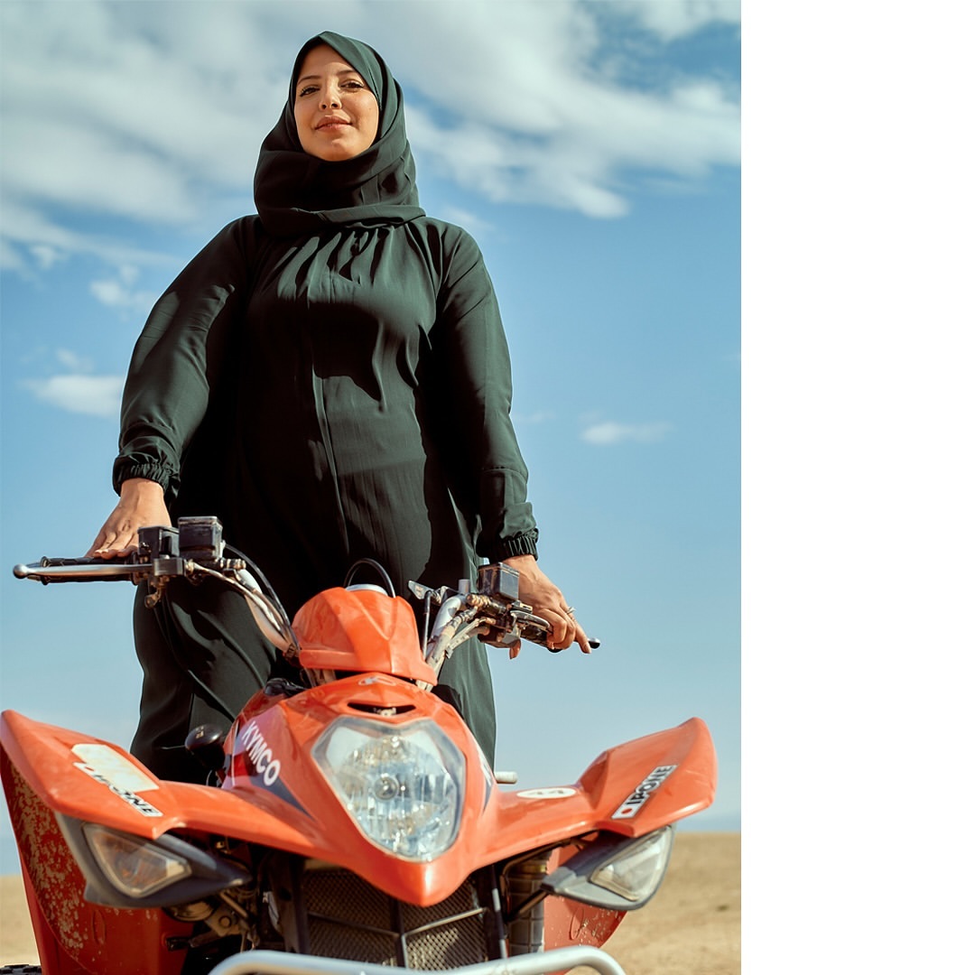 Adult woman in hijab sits on an orange all-terrain vehicle in a desert landscape with blue sky and clouds in the background.