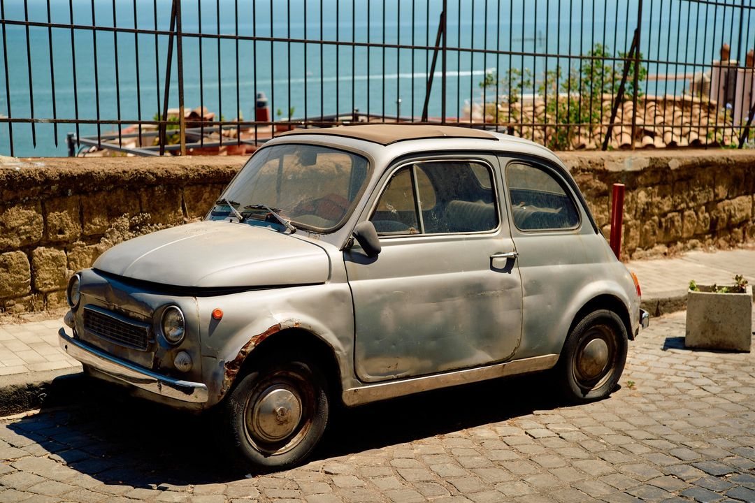 Weathered silver Fiat 500 parked on cobblestone street near stone wall and metal fence overlooking the ocean on a sunny day.