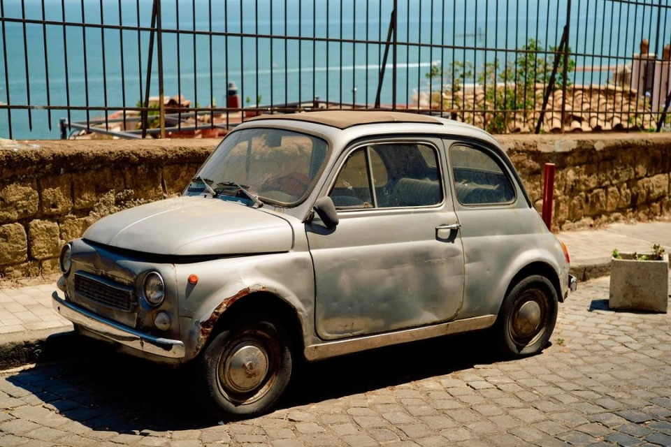 Weathered silver Fiat 500 parked on cobblestone street near stone wall and metal fence overlooking the ocean on a sunny day.