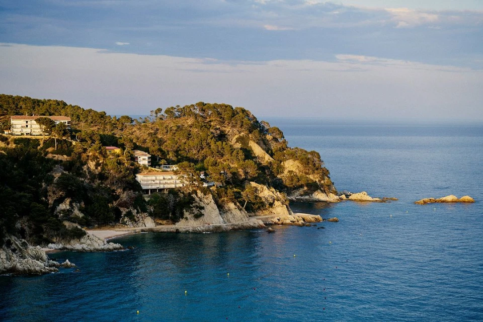Scenic view of ocean and coastline with trees and buildings under a cloudy sky, capturing nature's beauty.